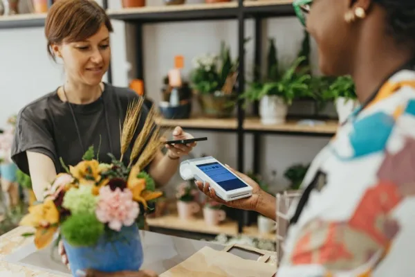 A customer is paying for a floral arrangement at a flower shop counter by tapping her smartphone to a point-of-sale device held by the cashier for a contactless transaction.